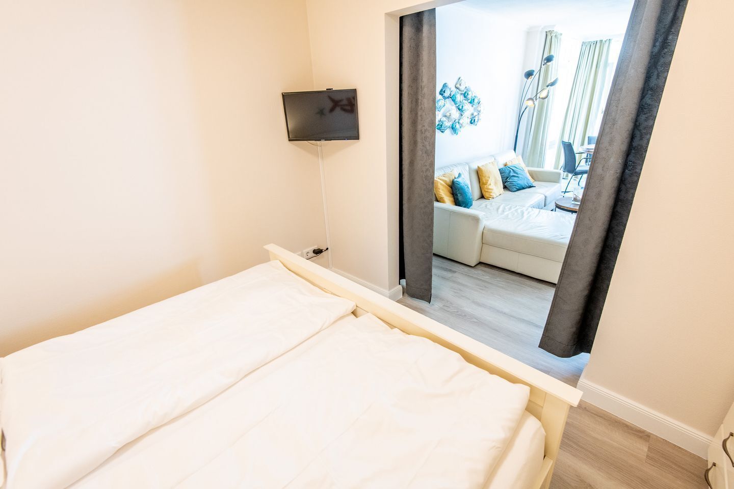 Un dormitorio con cama, televisor montado en la pared y vista a un salón con sofá y cortinas.