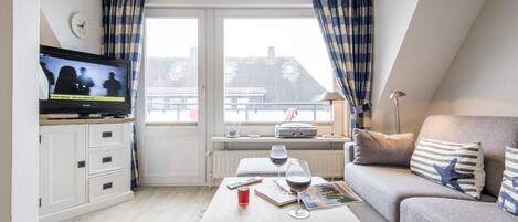 A cozy living room with a television, a sofa, a coffee table with drinks, and a view of the city through large windows.
