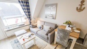 Un salon moderne avec un canapé, une table à manger et un petit bureau. Il y a une fenêtre avec des rideaux, un radiateur et un tableau représentant un voilier accroché au mur.