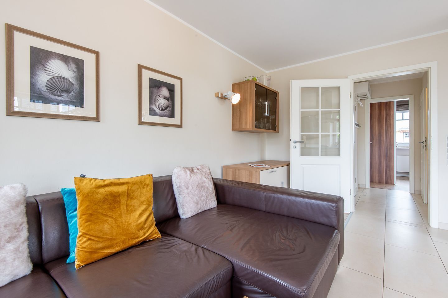 A modern living room with a brown leather sofa, two framed artworks, a wooden cabinet, and a white door leading to another room.