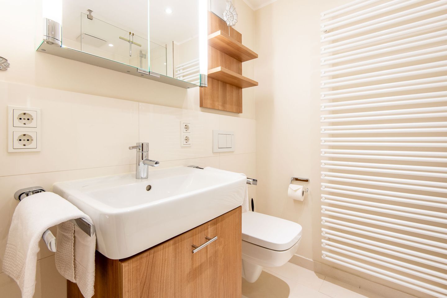 A modern bathroom with a white sink, wooden cabinet, and a toilet. There are two electrical outlets on the wall, a mirror above the sink, and a towel hanging on the side.