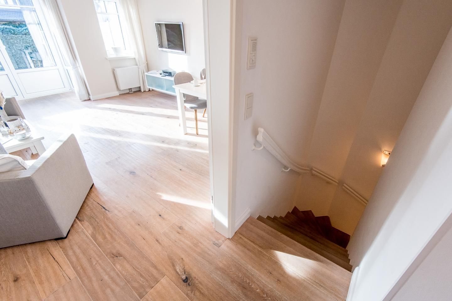 Un interior moderno con suelo de madera, una escalera blanca y una pequeña zona de estar con una mesa y sillas.