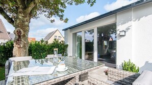 Ein Tisch mit Glasplatte, eine weiße Tasse und ein Untertasse, ein Korbstuhl und ein Baum im Hintergrund.