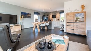 Ein modernes Wohnzimmer mit dunklem Boden, einem Holzespeisetisch mit Stühlen, einem Fernseher und einem großen Fenster.