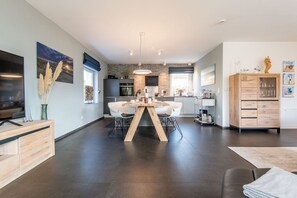 A modern kitchen with a brick backsplash, a dining area with a wooden table and white chairs, and a living room with a dark floor and a TV on a wooden stand.