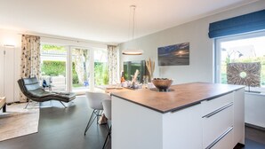 Eine moderne Küche mit einer großen Kochinsel, einem Essbereich mit einem Stuhl und Blick auf einen Garten durch große Fenster.