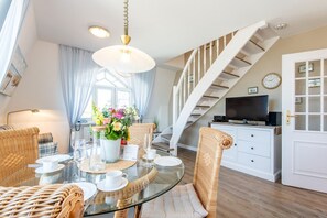 Ein Essbereich mit einem Glastisch, Korbstühlen und einer Kristallleuchter. Eine Treppe mit weißer Geländer führt eine weitere Ebene hinauf.