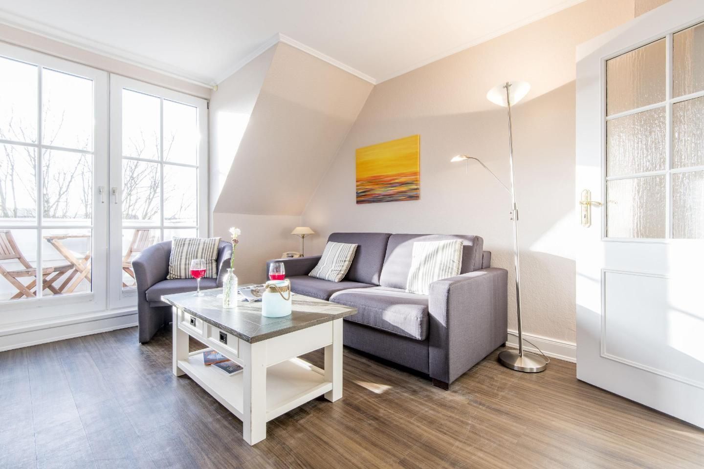 Un salon moderne avec un canapé gris, une table basse blanche et un lampadaire. Une grande fenêtre laisse entrer la lumière naturelle et offre une vue sur le mobilier d’extérieur.