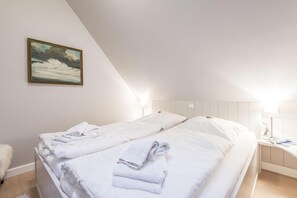 Une chambre à coucher avec deux lits, du linge de lit blanc, un tableau représentant une mer déchaînée et une table de chevet avec une lampe.