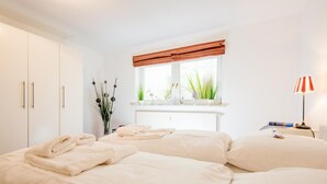 A bedroom with a large bed, a window with a view of greenery, and a bedside table with a lamp. - Old town gate in Friesenpark, apartment 7 (Sylt)