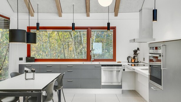 A modern kitchen with grey cabinets, a white countertop, and stainless steel appliances. There is a dining area with a table and chairs, and a window with a view of trees.
