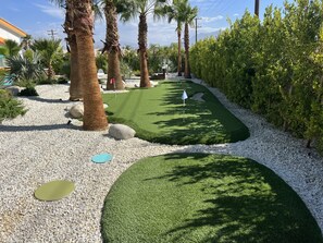 A well-maintained garden with palm trees, a golf putting area, and a pathway lined with white pebbles. - PS Playground - Putt Chip - Horseshoe - Corn Hole - Pool Table (Palm Springs)