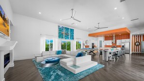 A modern living room with a sectional sofa, a dining area with a kitchen in the background, and a large window providing natural light. - PS Playground - Putt Chip - Horseshoe - Corn Hole - Pool Table (Palm Springs)