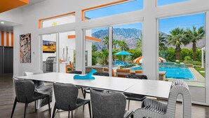 A modern dining area with a large table and chairs, large windows with mountain views, and a pool area visible through the windows. - PS Playground - Putt Chip - Horseshoe - Corn Hole - Pool Table (Palm Springs)