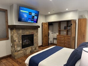 Ein Schlafzimmer mit Kamin aus Stein, einem Bett mit blauer und weißer Bettdecke, einem Fernseher, der über dem Kamin angebracht ist, und einem Holzschrank mit Spiegel.