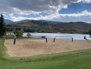Ein Beachvolleyballfeld mit Netz, umgeben von Sand und Gras, mit Blick auf einen See und Berge im Hintergrund.