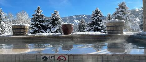 Una piscina con un marcador de profundidad de 9 metros y un letrero de no buceo, rodeada de árboles cubiertos de nieve y una montaña al fondo.