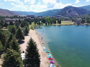Un complexe hôtelier au bord d’un lac, doté d’une plage de sable, de rangées de kayaks aux couleurs vives, et offrant en toile de fond un paysage de montagnes et de bâtiments.