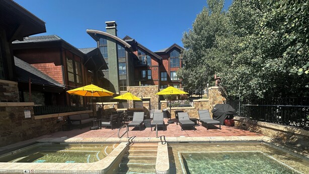 A clear pool with lounge chairs and umbrellas, surrounded by stone walls and a building with large windows.