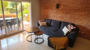 Un salon avec un canapé bleu foncé, une table basse ronde et une petite table d’appoint. Un mur en briques se trouve derrière le canapé, et une porte-fenêtre coulissante donne accès à un espace extérieur aménagé avec une table et des chaises.
