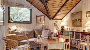 Um quarto acolhedor com teto de madeira, um sofá, uma mesa de jantar com cadeiras, uma estante de livros e uma janela com vista para árvores.