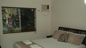 A neatly made bed with a checkered bedspread and two pillows, a window with a view of greenery, and a wall-mounted air conditioner. - Beachfront  (Yorkeys Knob)