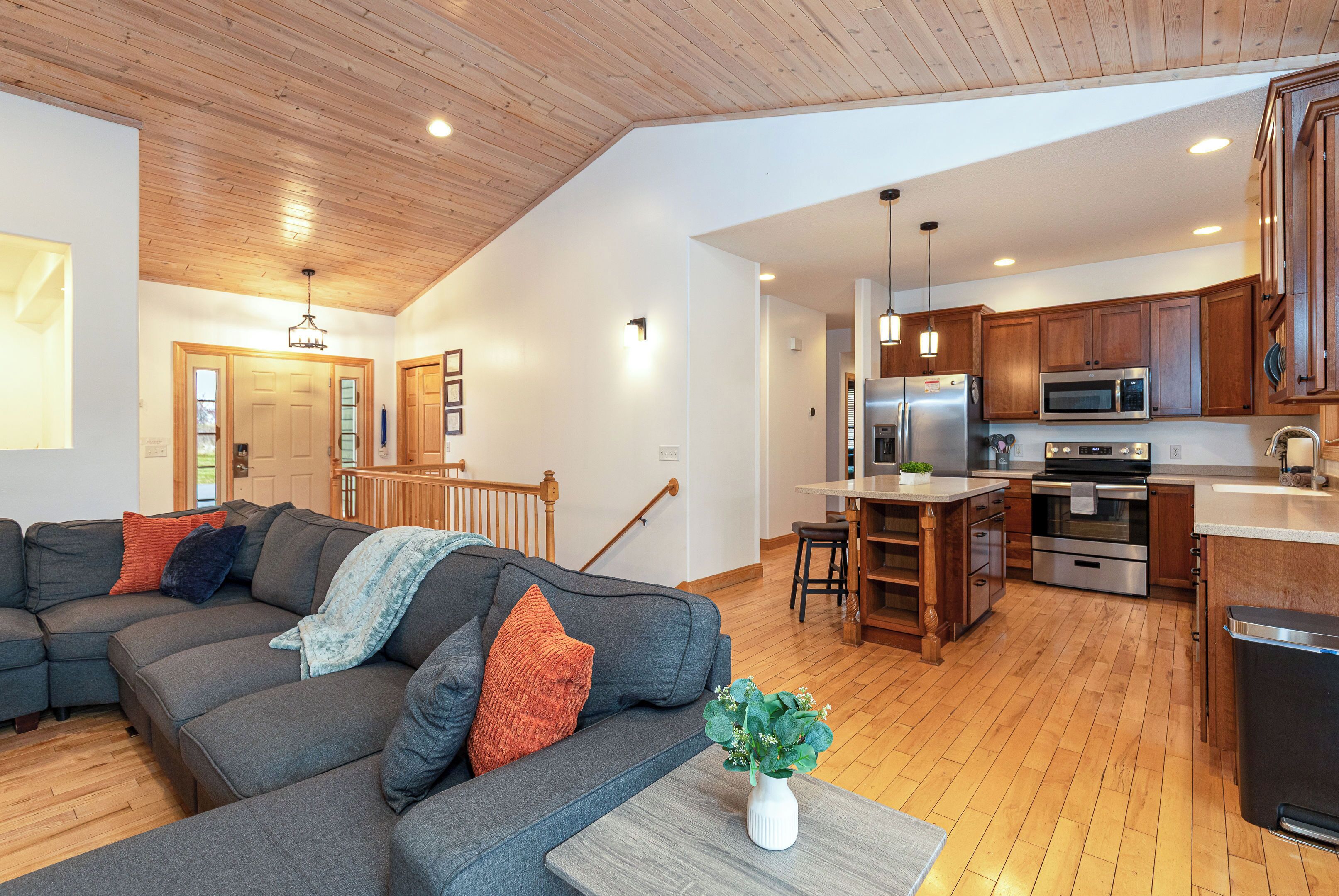 A modern living room with a grey sectional sofa, wooden ceiling, and a kitchen area with stainless steel appliances.