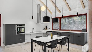 Eine moderne Küche mit Einbauofen, einem Esstisch mit Stühlen, Pendelleuchten und einem großen Fenster mit Blick auf Bäume.