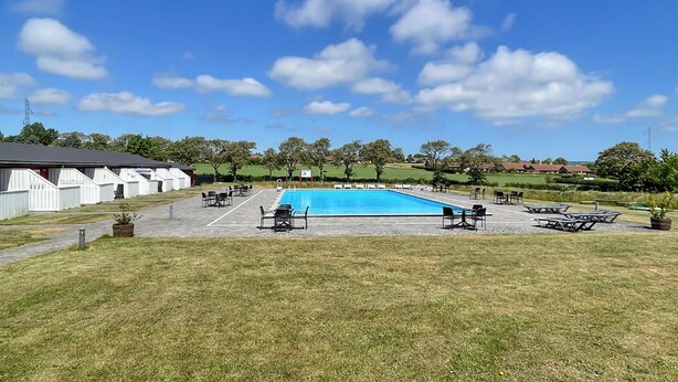 Una piscina con lettini e tavoli, circondata da un prato e un edificio con tetto spiovente.