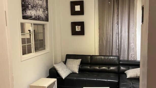 Una sala de estar con un sofá de cuero negro, dos cuadros en la pared, una ventana y una cortina.