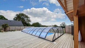 Un patio couvert doté d’un toit transparent, d’une terrasse en bois et offrant une vue sur une maison en arrière-plan.
