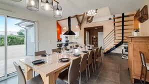 Un espace repas avec une longue table dressée pour le repas, une cuisine en arrière-plan et un escalier menant à un niveau supérieur.