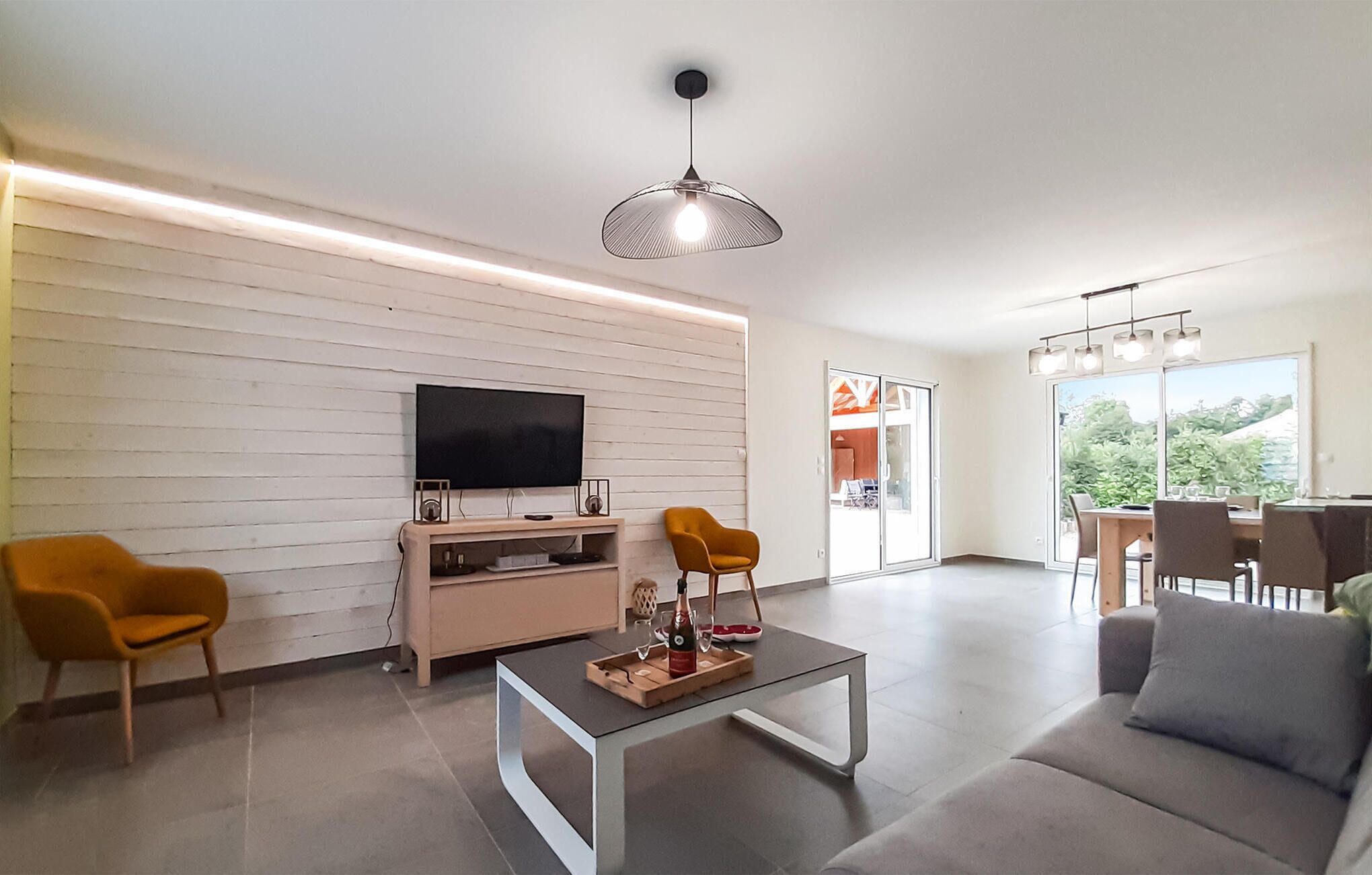 A modern living room with a flat-screen TV, a coffee table with a bottle and glasses, two armchairs, and a dining area with a table and chairs.