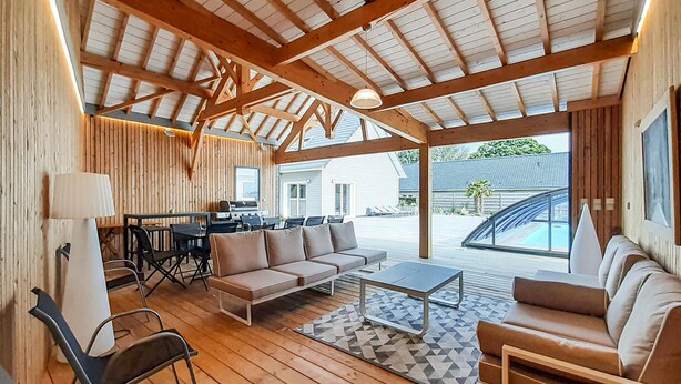 Une vaste terrasse dotée d’un plafond en bois, un coin repas avec un barbecue et un espace détente avec un canapé et des fauteuils.