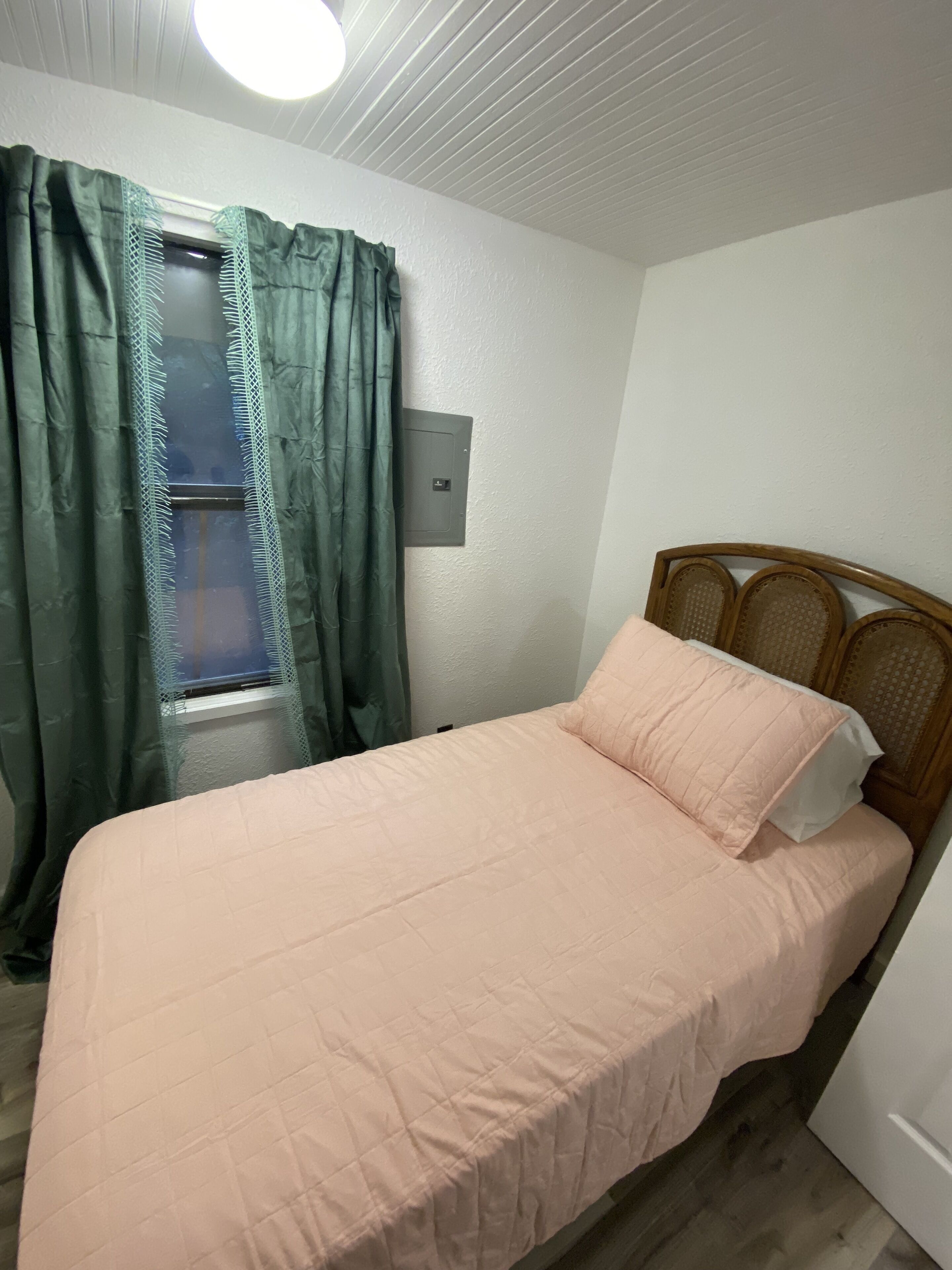 A small, neatly made bed with a wooden headboard, a window with curtains, and a wall-mounted light fixture.