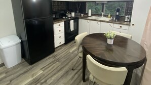 Une cuisine de taille modeste, avec des meubles blancs, un réfrigérateur noir, une table à manger en bois accompagnée de chaises, et une fenêtre donnant sur un paysage verdoyant.