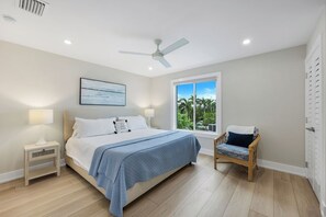 A bedroom with a bed, bedside tables, a chair, a ceiling fan, a window with a view of greenery, and a framed picture on the wall.