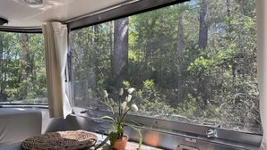 A cozy interior with a large window offering a forest view, a woven pouf, and a potted plant. - Large Home w 2 Airstream Glampers & Spa near Beach & Downtown (Savannah)