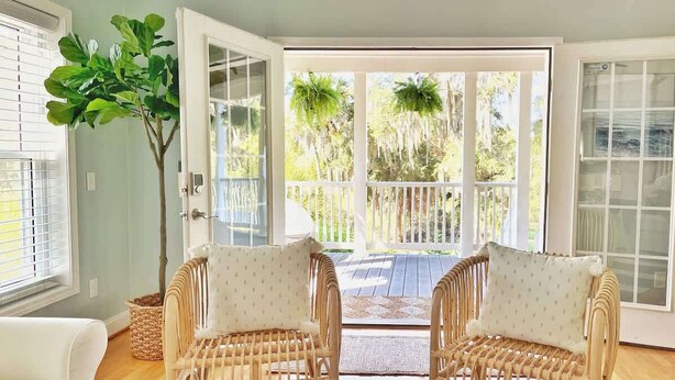 A room with two wicker chairs, a large potted plant, and a view of a balcony with outdoor furniture. - Large Home w 2 Airstream Glampers & Spa near Beach & Downtown (Savannah)