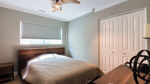 Un dormitorio con una cama, una mesita de noche de madera, un escritorio con una silla, un ventilador de techo y una ventana con persianas.