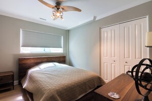 Una camera da letto con un letto, un comodino in legno, una scrivania con una sedia, una ventola a soffitto e una finestra con persiane.