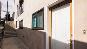 Un edificio con una puerta blanca marcada con la letra 'E', un conjunto de escaleras que conducen a un rellano, y una ventana con persianas.