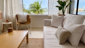 Un salon moderne avec un canapé blanc, un fauteuil en osier, une petite table ronde et une grande fenêtre donnant sur des palmiers et un immeuble.