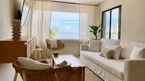 Un salon avec un canapé blanc, des fauteuils en osier, une table basse, un ventilateur de plafond, une grande fenêtre donnant sur la mer et une plante.