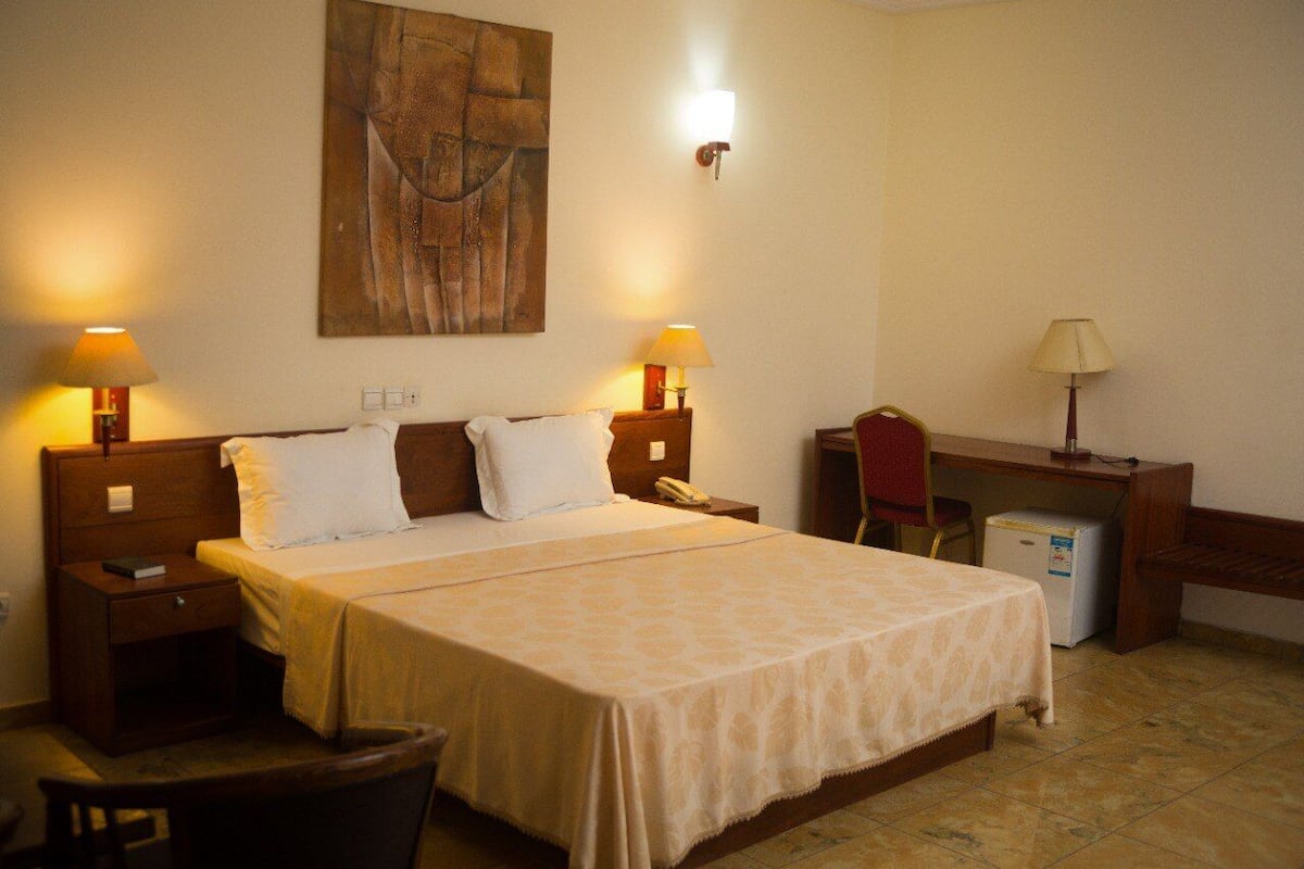 A hotel room with a bed, two bedside lamps, a desk, a chair, and a refrigerator.