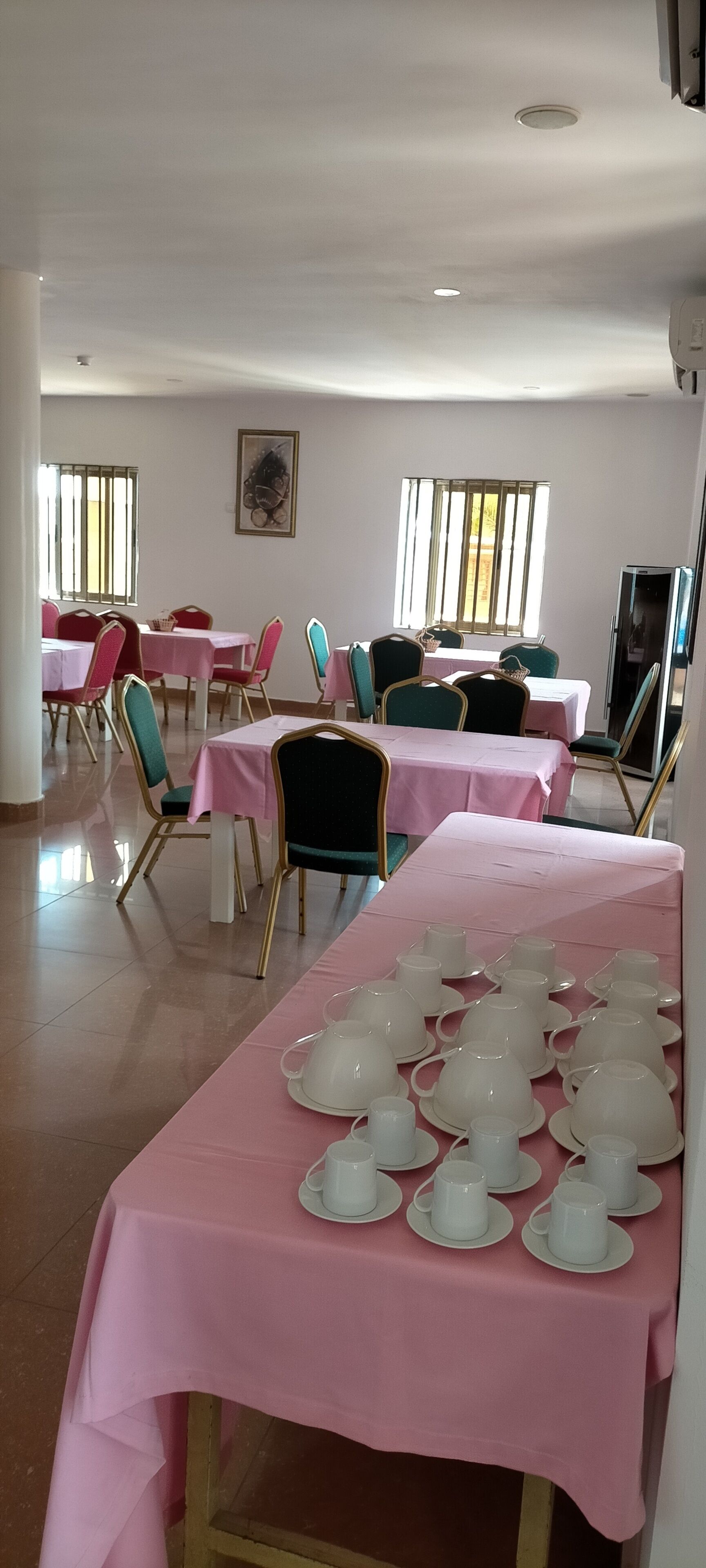 A dining area with pink tablecloths and chairs, a long table with teacups and saucers, and a refrigerator in the background.