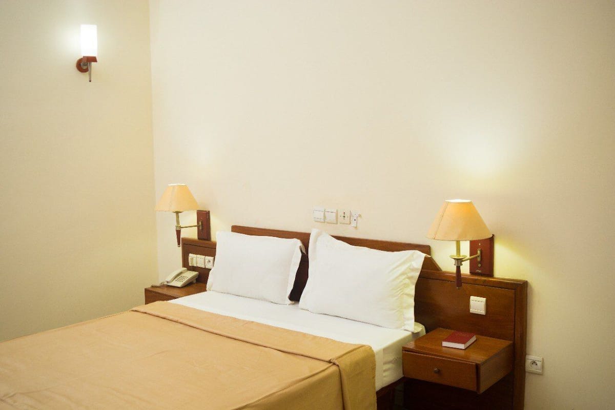 A hotel room with a bed, two bedside lamps, a bedside telephone, and a small bedside table with a book.