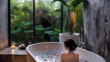 A bathtub with rose petals, a candle, and a potted plant by a window.