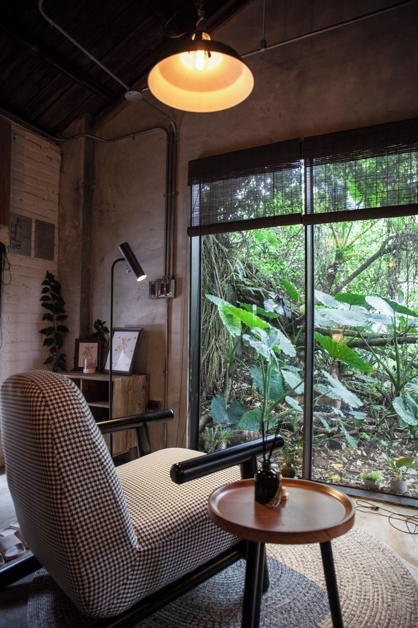 Une pièce chaleureuse avec un fauteuil à carreaux, une table ronde en bois, un luminaire suspendu et une grande fenêtre donnant sur un paysage verdoyant.