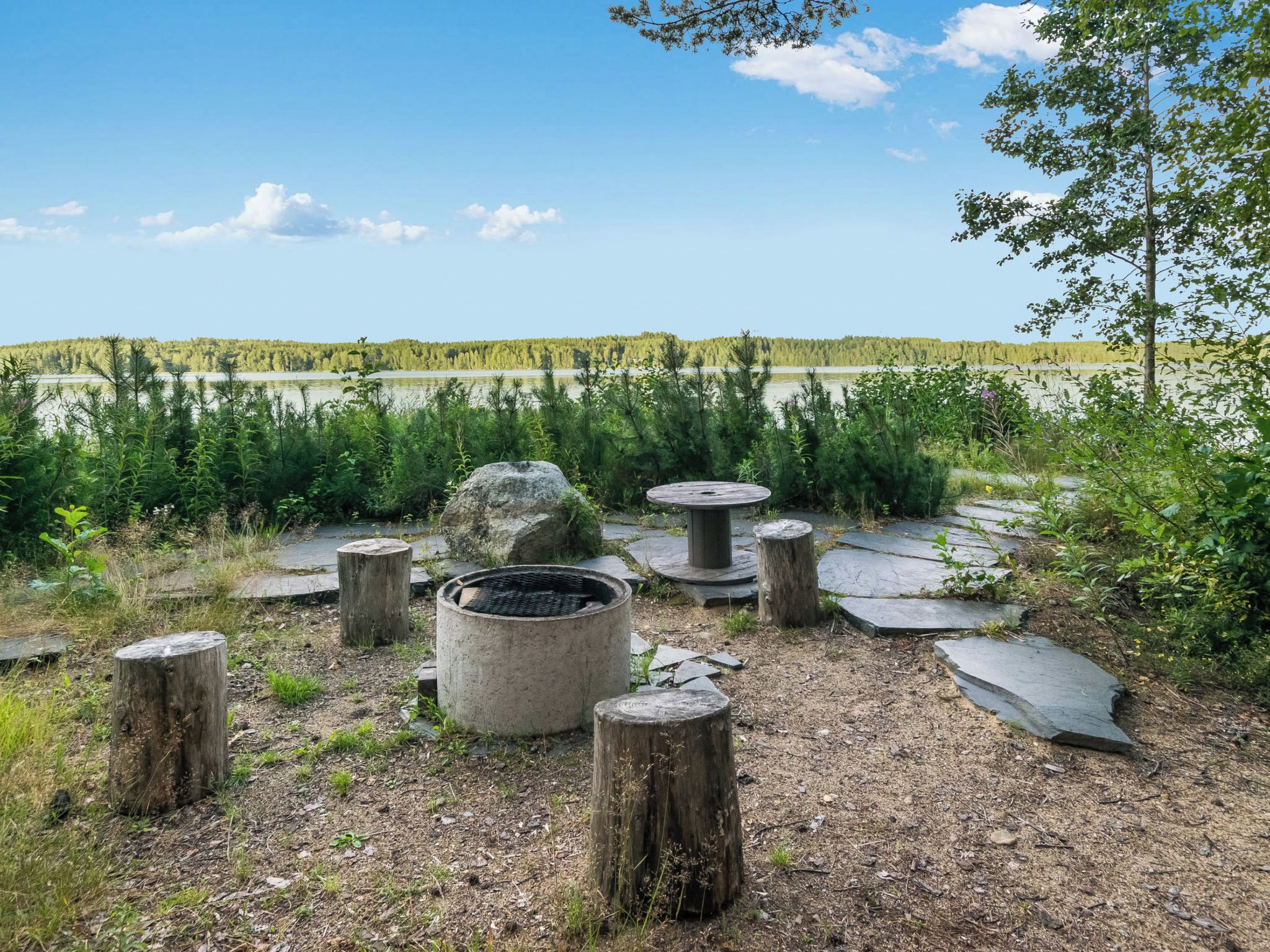 Een buitengebied met een vuurkuil, stenen banken en uitzicht op een meer en bos.